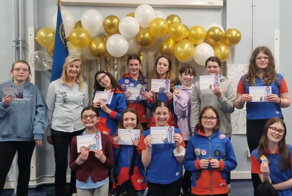 A group of Girl Guides holding up their award certificates, alongside their leaders. Gold and white balloons are floating behind their heads