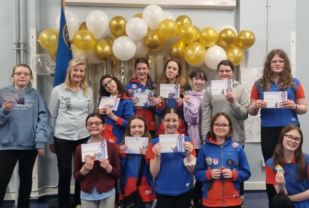 A group of Girl Guides holding up their award certificates, alongside their leaders. Gold and white balloons are floating behind their heads