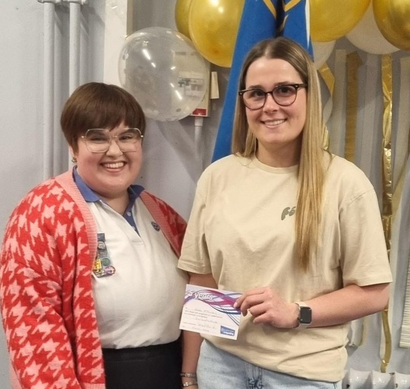 Katie stands alongside Greta, who is holding her certificate, with balloons in the background
