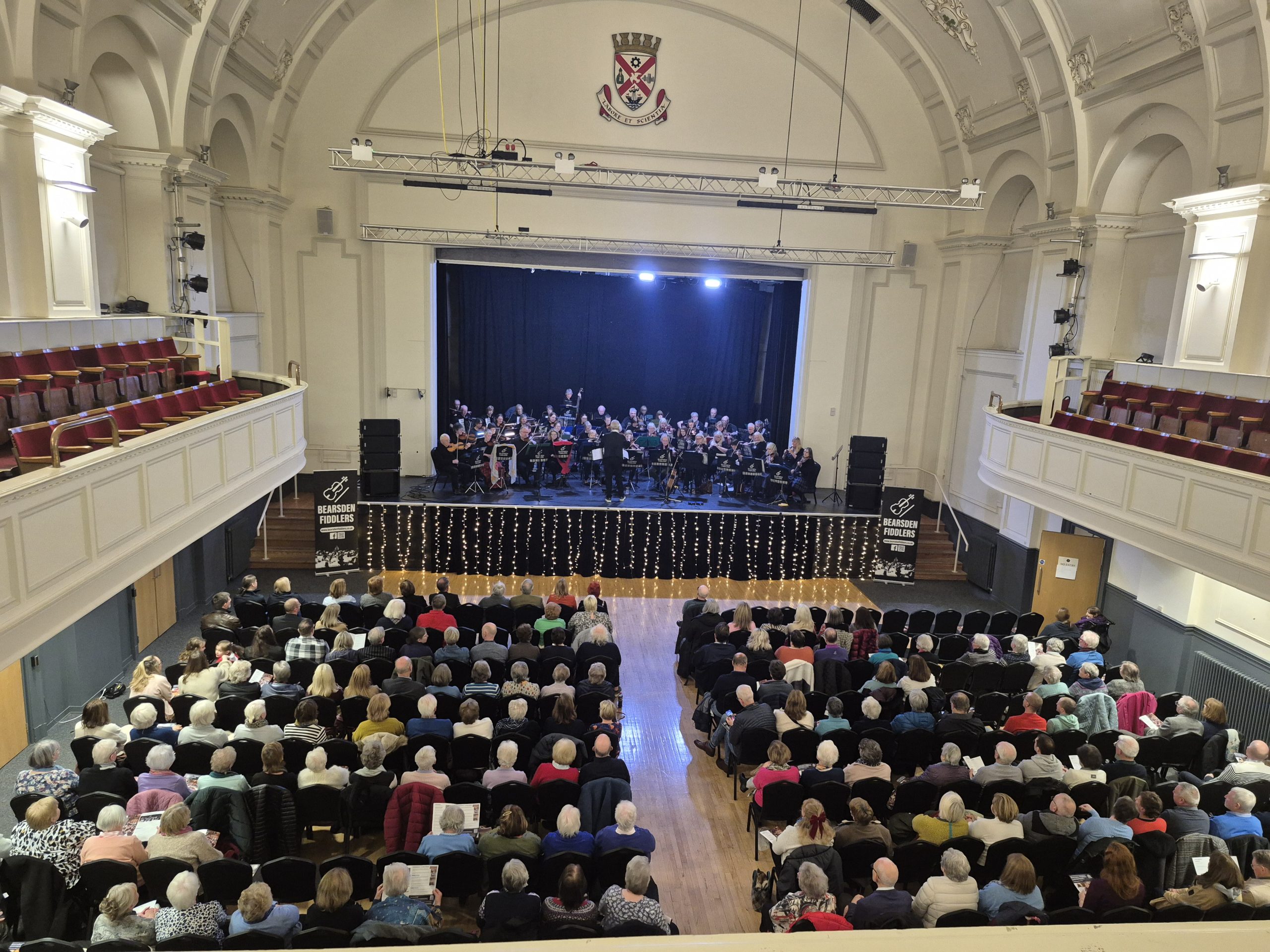 Clydebank Town Hall with a large audience seated facing a stage. Bearsden Fiddlers perform under bright lights, with an elevated balcony and arched ceiling visible.
