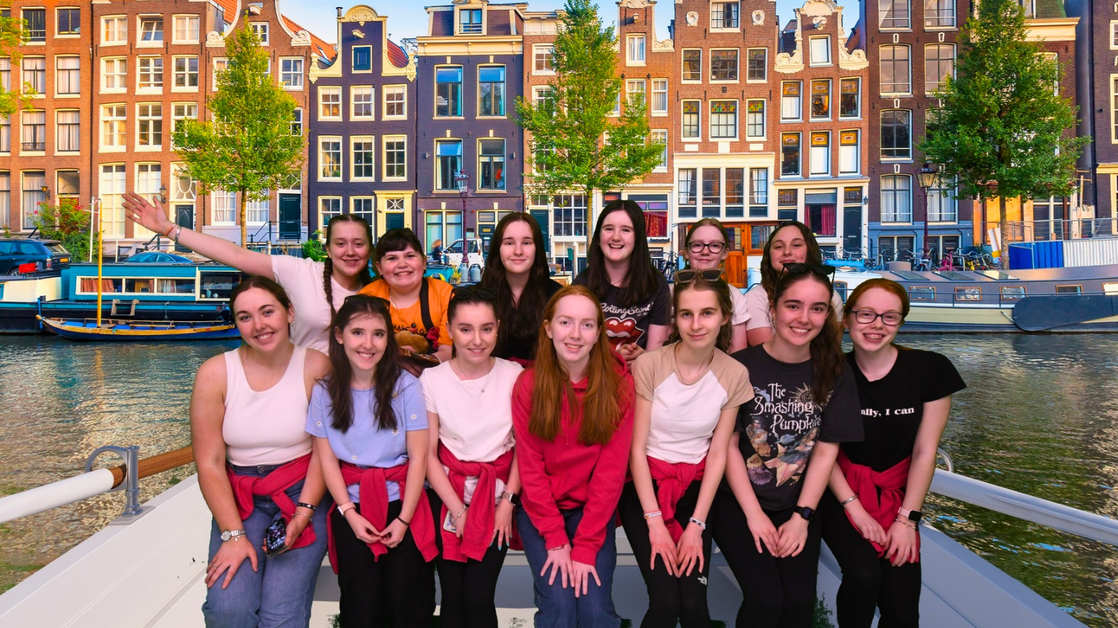 A group of fourteen smiling girls and young women, members of the 6th Clydebank Guides and Rangers, pose together on a boat in front of a scenic Amsterdam canal. Behind them are traditional Dutch buildings and houseboats, with trees lining the water. Some of the girls are wearing matching red hoodies or sweatshirts tied around their waists, and a few wear Guiding-themed shirts. The mood is cheerful and celebratory.