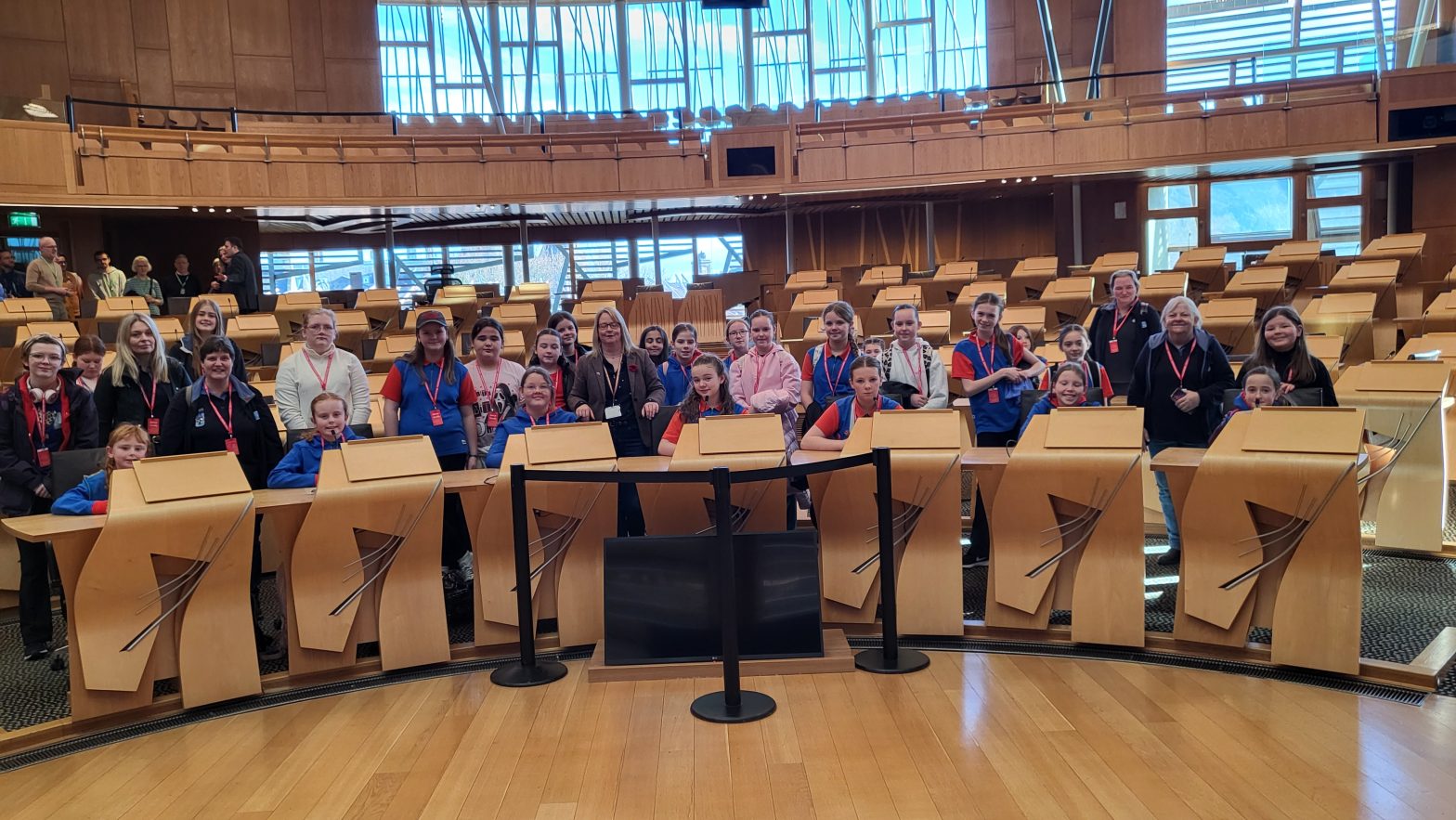 A large group of Girlguiding members—mostly girls in blue and red uniforms along with several adult leaders—stand together inside the debating chamber of the Scottish Parliament. They are positioned behind the curved wooden desks, smiling at the camera. The chamber features distinctive modern wooden architecture, tiered seating, and large windows letting in natural light.