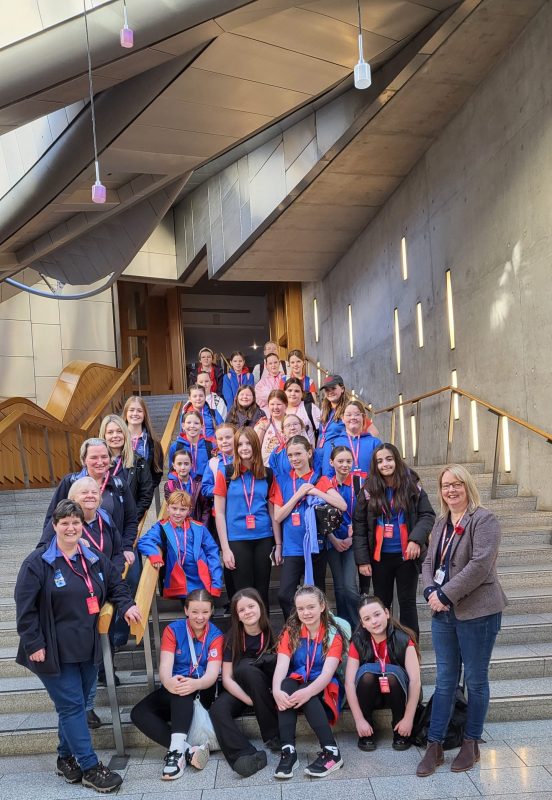 A large group of Girlguiding members—girls in blue and red uniforms along with several adult leaders—pose together on a wide staircase inside the Scottish Parliament building. They are smiling and wearing visitor lanyards. The modern interior features concrete walls with vertical light panels, wooden handrails, and geometric ceiling structures. Marie McNair MSP stands to the right of the group at the bottom of the stairs