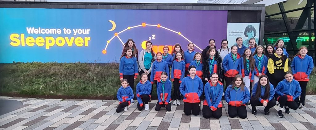 Guides in red and blue uniform outside the science centre, with a digital sign that reads: Welcome to your sleepover