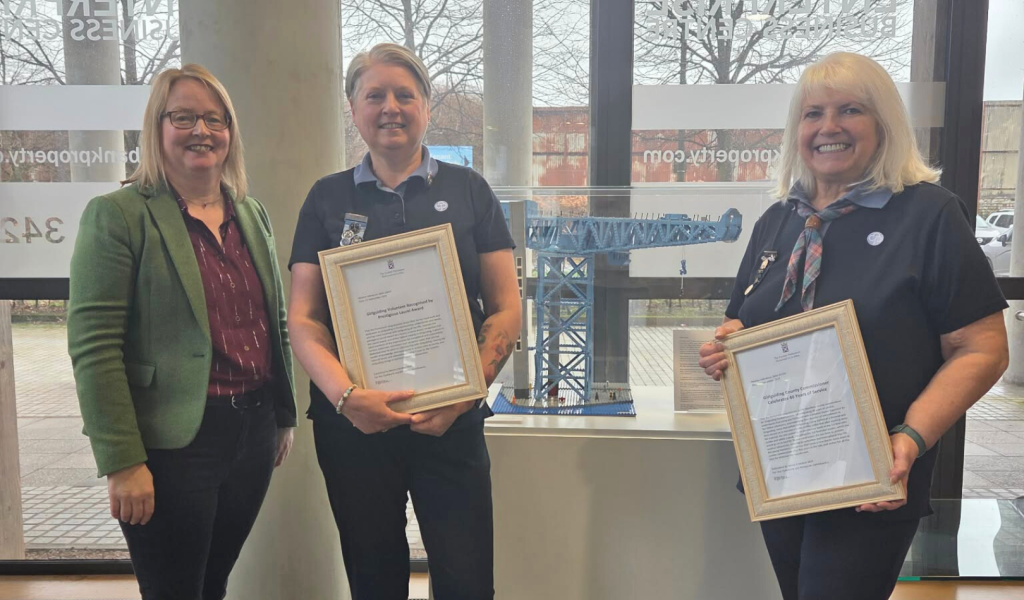 Marie McNair MSP, Janice Blair and Lorna Hamilton stand indoors in front of large windows, smiling at the camera. Janice and Lorna wear navy Girlguiding uniforms and hold framed certificates, while Marie wears a green blazer and dark trousers. A model crane is displayed on a pedestal behind them, with leafless trees visible outside.