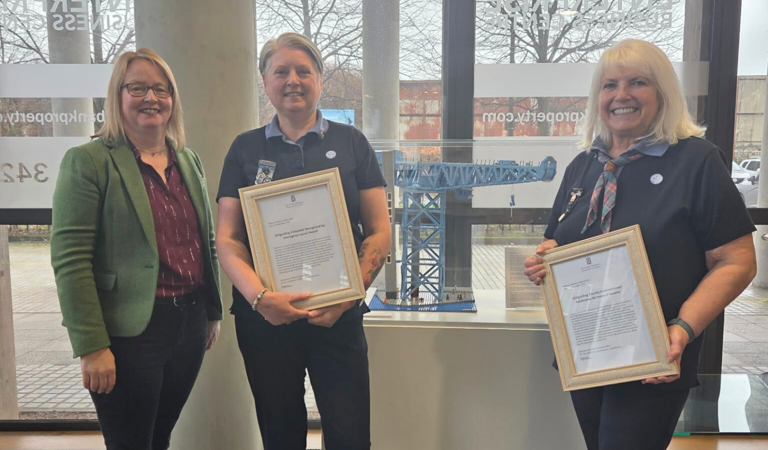 Marie McNair MSP, Janice Blair and Lorna Hamilton stand indoors in front of large windows, smiling at the camera. Janice and Lorna wear navy Girlguiding uniforms and hold framed certificates, while Marie wears a green blazer and dark trousers. A model crane is displayed on a pedestal behind them, with leafless trees visible outside.
