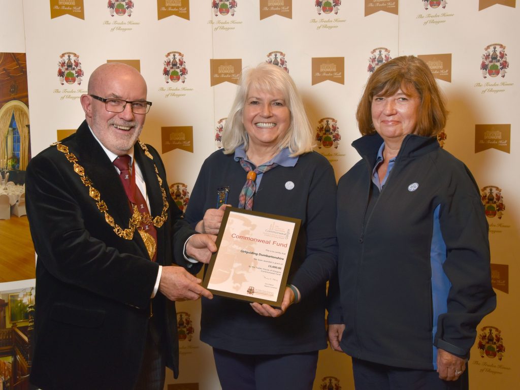 Rev Tom Pollock, Deacon Convener of the Trades House of Glasgow, presents a framed Commonweal Fund certificate to Lorna Hamilton, outgoing County Commissioner of Girlguiding Dunbartonshire, who is holding the award. Lynne Gibbons, the new County Commissioner, stands beside them. All three are smiling and posing in front of a Trades House of Glasgow branded backdrop during the presentation of the £5,000 funding award.