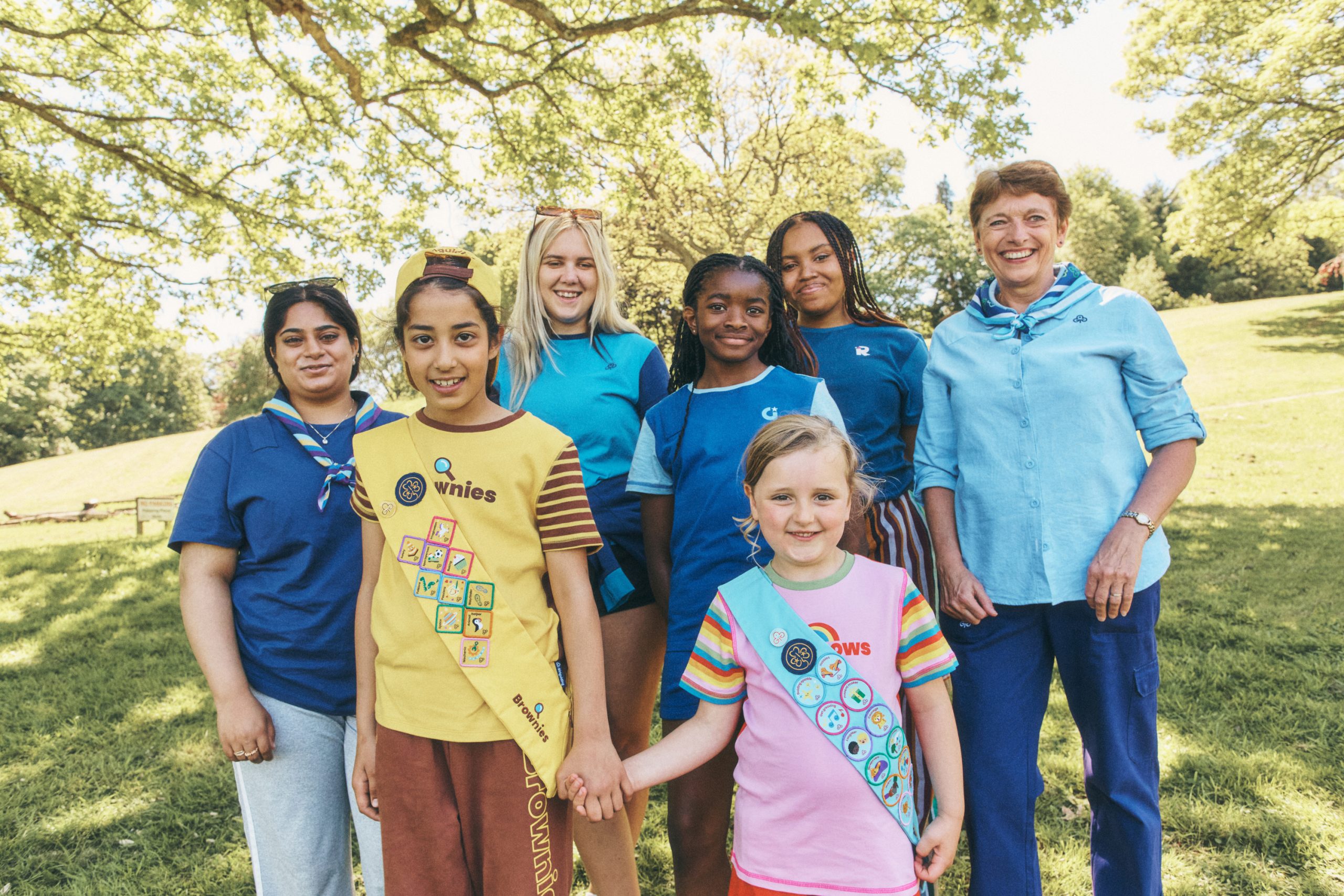 Group photo of seven people standing together on a grassy lawn in a park beneath large leafy trees. Two children stand at the front, holding hands. One child wears a yellow Brownies uniform with several achievement badges, and the other wears a pink top with a light-blue sash covered in badges. Behind them are five older children and adults wearing blue uniform-style tops and scarves. The scene is outdoors on a sunny day, with dappled sunlight filtering through the tree canopy and open green space in the background.