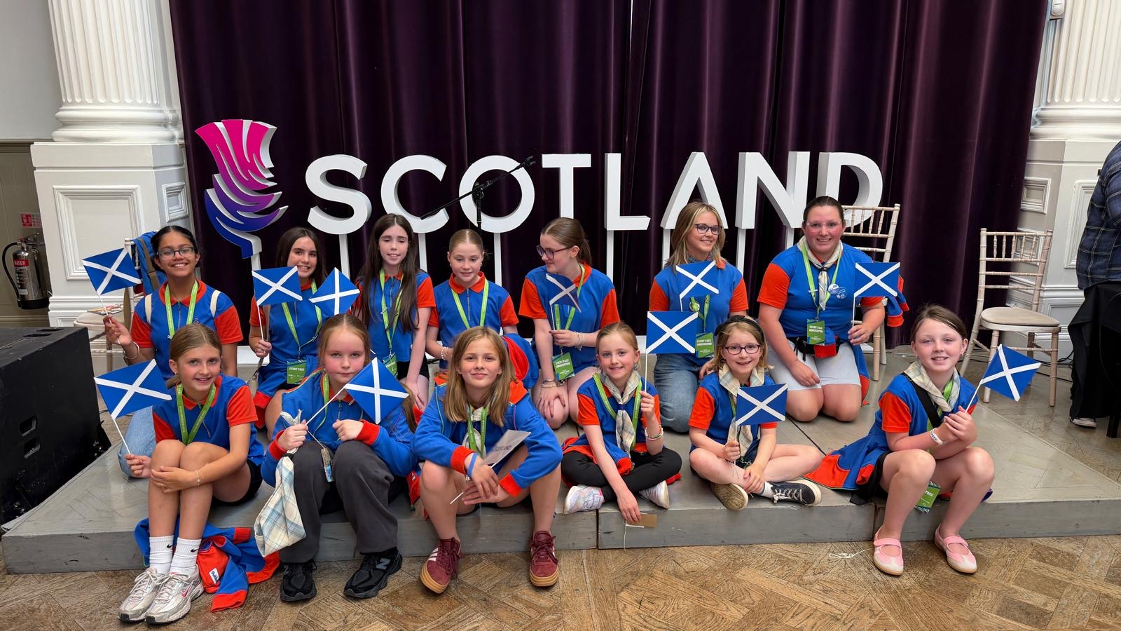 Group photo of approximately 12 Girl Guides in blue and red uniforms sitting and kneeling on a small stage, each holding a Scottish Saltire flag. Behind the group is a large white “SCOTLAND” sign with a purple-and-pink emblem, set against dark purple curtains. The photo is taken inside an elegant hall with white columns, decorative wall panels, and wooden flooring. Several chairs and a microphone stand are visible in the background.