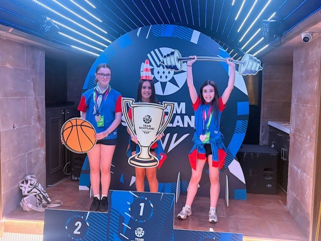 Three Girl Guides wearing blue and red team uniform, event lanyards, and medals pose on a winners’ podium in front of a large Team Scotland backdrop. The participant on the left stands on the second-place platform beside a large basketball prop. The participant in the centre stands on the first-place platform holding a large Team Scotland trophy cut-out and wearing a traffic cone hat. The participant on the right stands beside the podium holding a barbell prop overhead. The venue features blue illuminated ceiling lights, wood-patterned flooring, and stone-effect walls, creating a playful photo opportunity area.