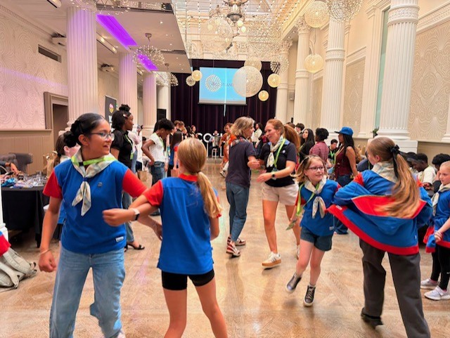 Several Girl Guides in blue uniforms with red sleeves and neck scarves are gathered on a polished dance floor in an ornate hall. Some are dancing and holding hands in the foreground, while others are standing and talking throughout the room. The hall features tall white columns, decorative walls, large hanging light fixtures, and purple accent lighting. At the far end of the room, a presentation screen displays a Team Scotland logo above a stage area. Tables and additional attendees line the sides of the hall, creating a lively event atmosphere.