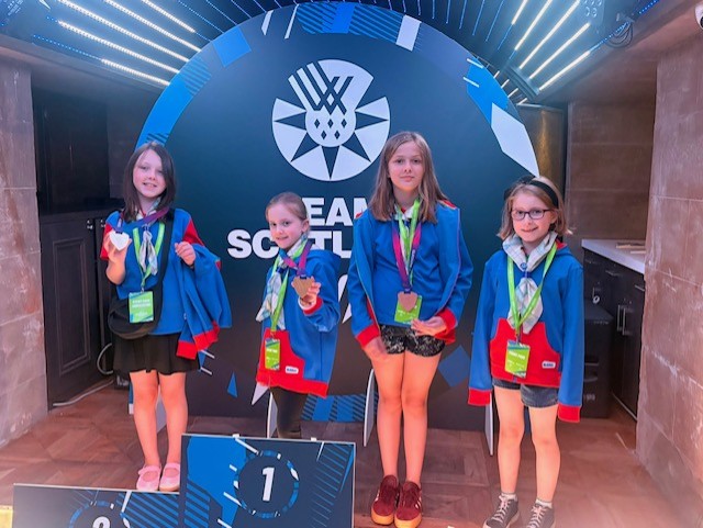Four young Girl Guides wearing blue jackets with red cuffs and green event lanyards stand in front of a large circular backdrop displaying the Team Scotland logo and the words “TEAM SCOTLAND.” Each participant is wearing a medal and holding part of the jacket open to show it. A winners’ podium with the number “1” is visible in the foreground. The photo is taken indoors in a brightly lit venue with illuminated ceiling panels and wood-patterned flooring.