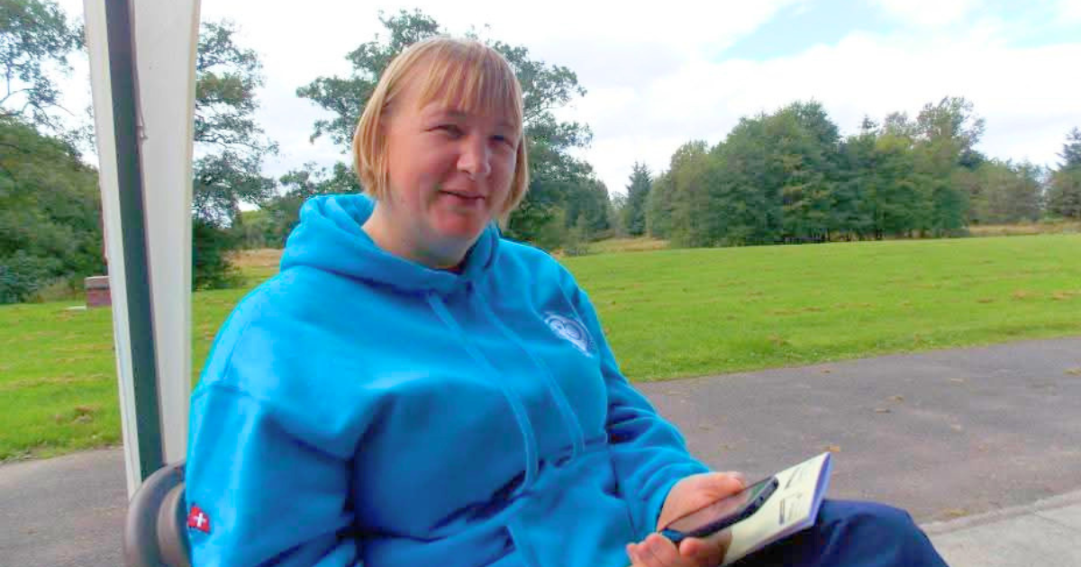 Carol-Ann sitting outdoors on a chair, wearing a bright blue hoodie, holding a smartphone and a booklet. She is in a grassy park or outdoor activity area, with trees and a paved path in the background.