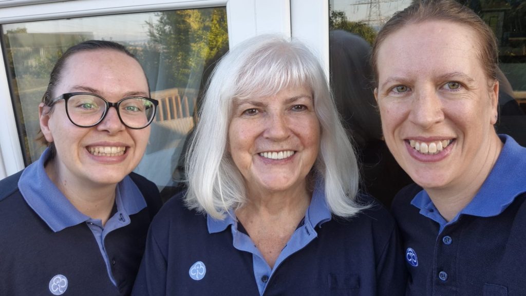 Three people - Cat, Lorna and Sarah - in matching navy-blue Girlguiding polo shirts pose for a selfie outdoors beside a glass door, with trees and reflections visible in the background.