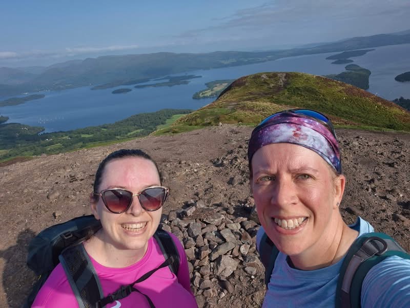 Cat and Sarah wearing backpacks take a selfie from a rocky hilltop, with a panoramic view of a large loch, wooded islands and rolling hills stretching into the distance under a clear sky.
